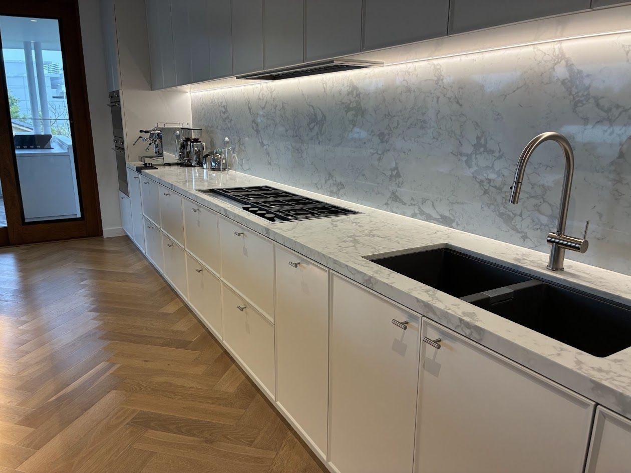 Long white kitchen with marble-look benchtop and lit splashback, black sink and herringbone timber floor
